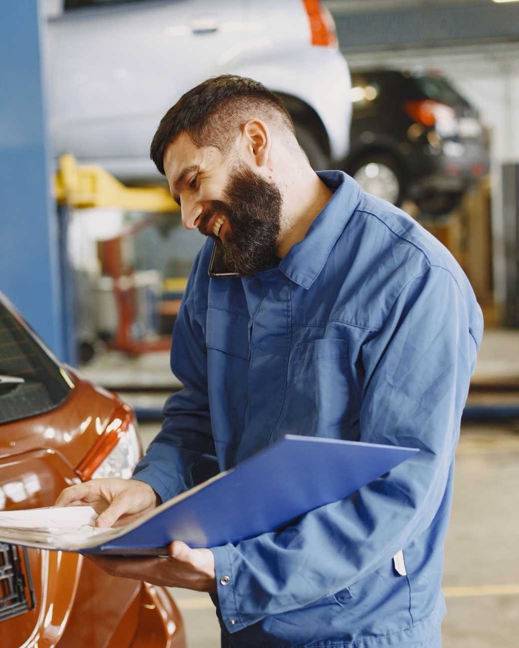 A smiling mechanic on the phone, holding a binder of work orders next to a car