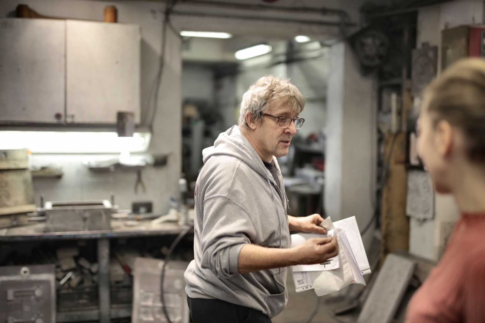 A man in a grey hoodie holds a stack of papers and talks with a coworker in a metal workshop.